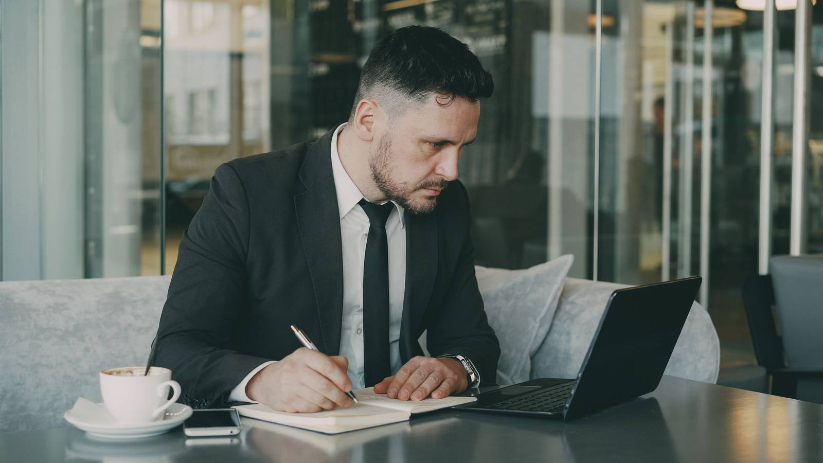 A man working on a laptop and taking notes at a table in a modern café.