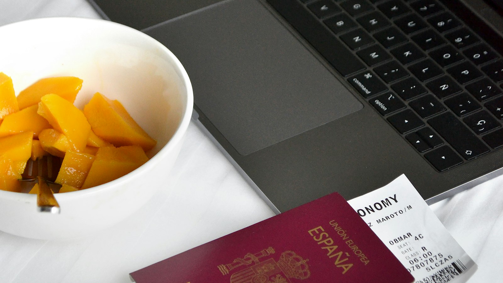 A passport and a boarding pass next to an open laptop, with a bowl of fruit alongside