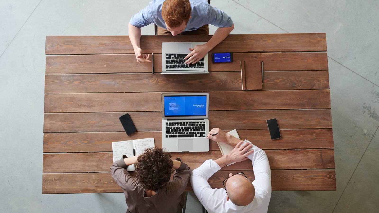 Three colleagues working with laptops and phones around a wooden table in an office.
